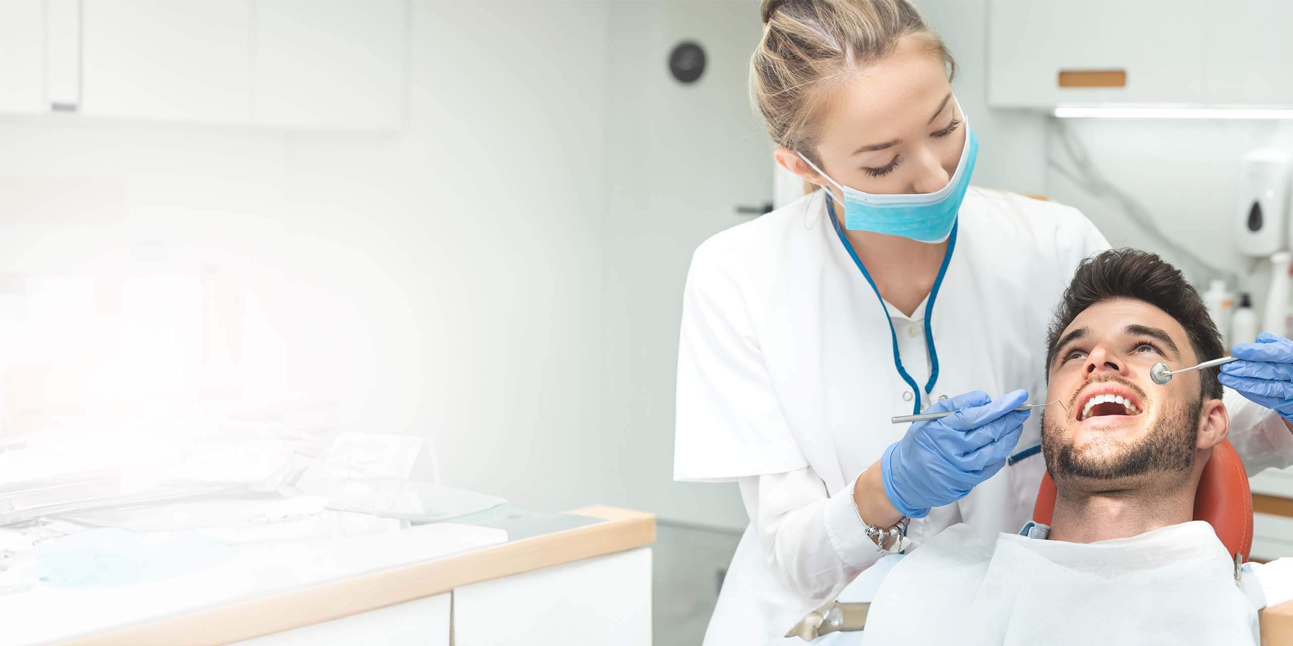 Ein junger männlicher Patient im Zahnarztstuhl wird in einer modernen Zahnklinik von einer Zahnärztin untersucht, die blaue Handschuhe und eine OP-Maske trägt.