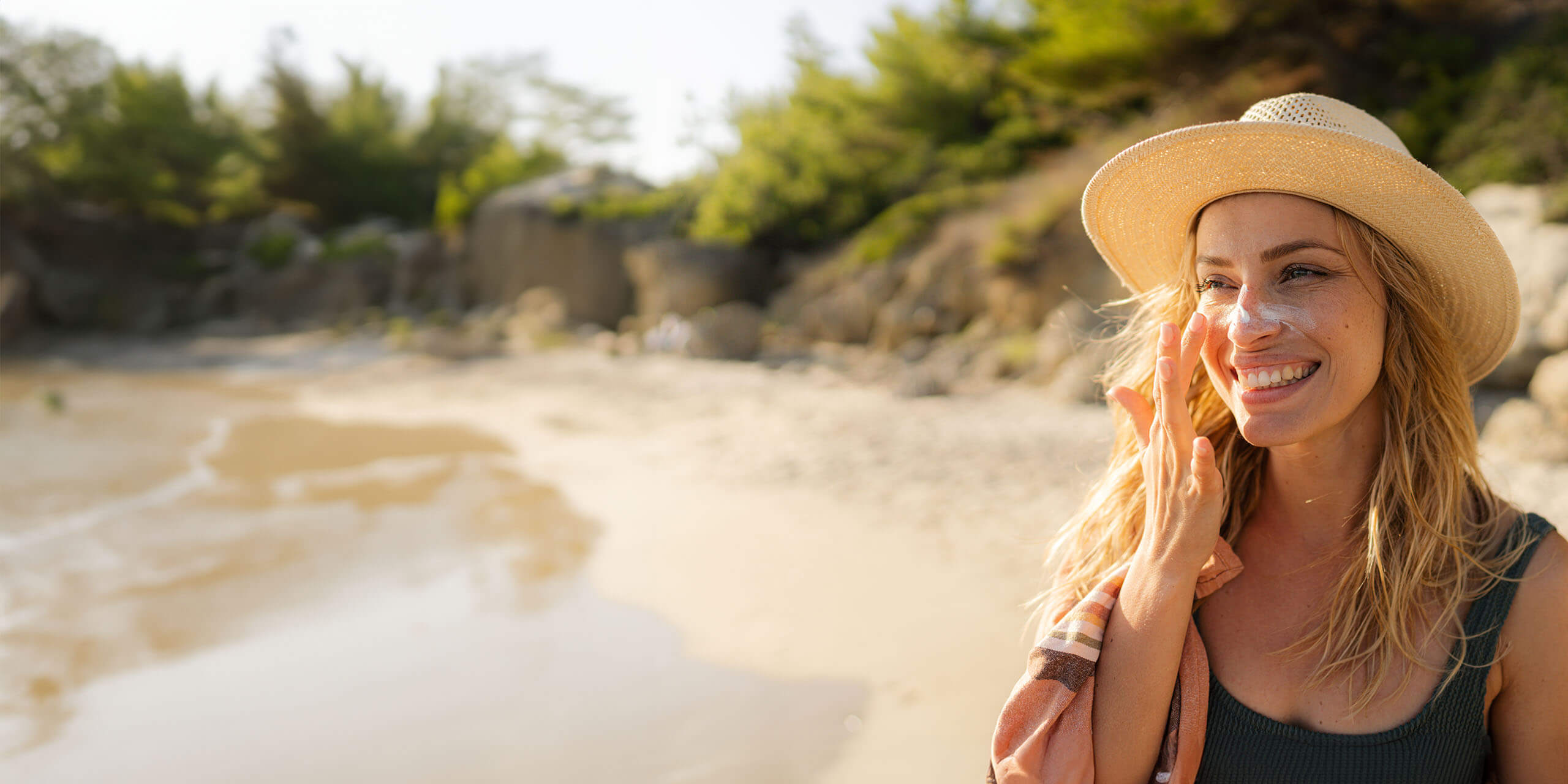 Eine lächelnde Frau mit Sonnenhut steht an einem Sandstrand und schützt ihre Augen mit der Hand vor der Sonne. Im Hintergrund plätschern sanft Wellen ans Ufer.