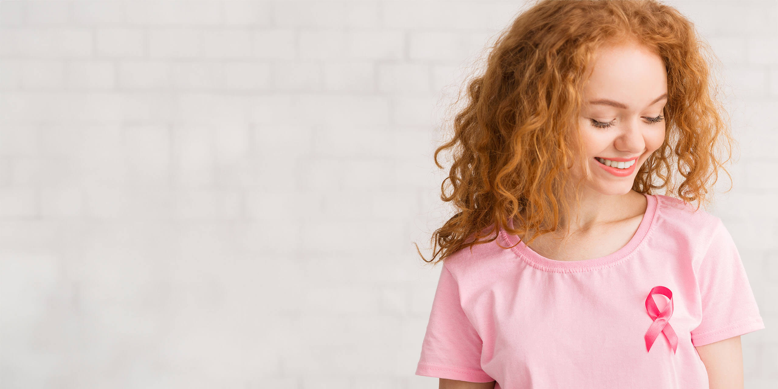 Eine junge Frau mit lockigem rotem Haar, einem rosa T-Shirt und einer rosa Schleife als Anstecknadel, lächelt vor einem weißen Ziegelhintergrund.